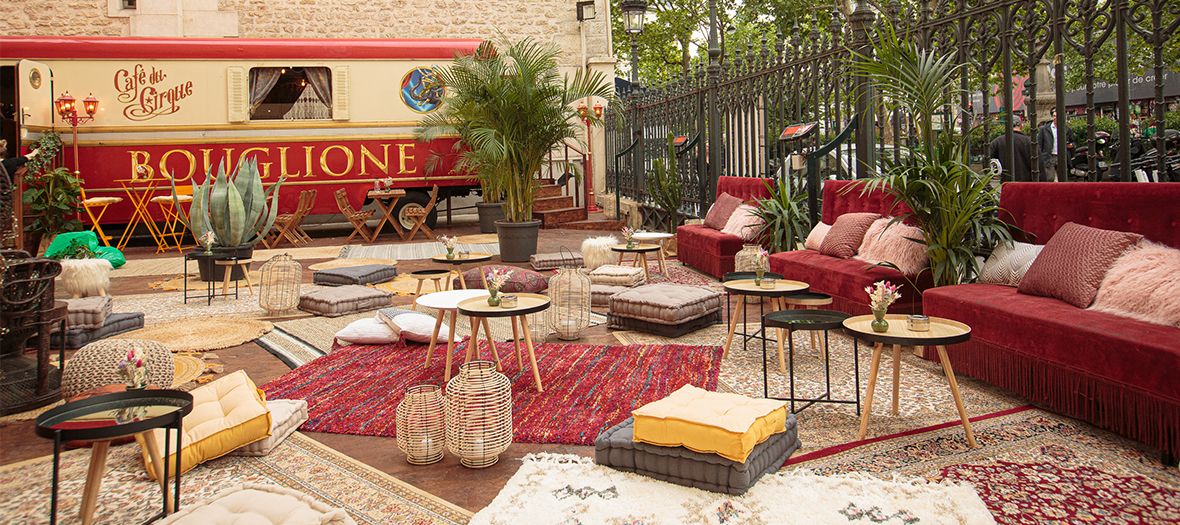 Terrasse éphémère du Gyspy café avec fauteuil Emmanuelle, banquettes crapauds, mix and match de tapis en jute, coussins douillets, poufs en tricot, plantes tropicales dans la cour du Cirque d'Hiver à Paris